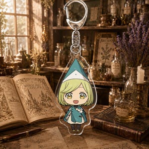 May include: A clear acrylic keychain featuring a stylized anime character with short blonde hair, large golden eyes, and a wide smile. The character wears a teal pointed hat and a matching teal cape over a white outfit, attached to a silver metal clasp.