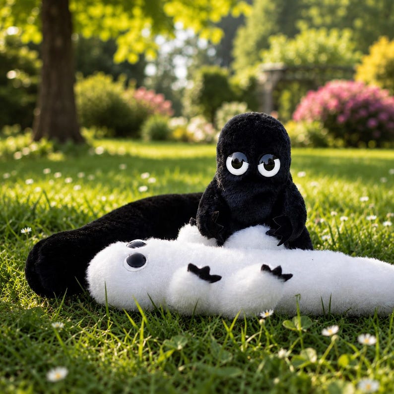 May include: A plush toy set featuring a black creature with large white eyes and a white creature with large black eyes. Both creatures have rounded bodies, short limbs, and soft textures, positioned together on a green grassy surface.