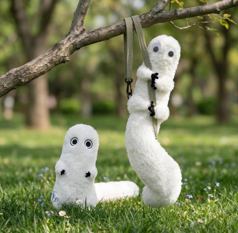 May include: Two white plush toys with soft, fuzzy textures, large round eyes with black pupils, and small black hands. One plush toy stands upright on the grass, while the other hangs from a tree branch by a light green fabric strap.