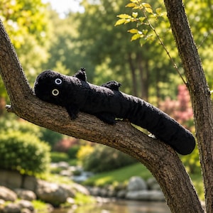 May include: A long, black plush toy shaped like a salamander or lizard with a soft, fuzzy texture. It features two prominent, circular white eyes with black pupils and four small, stubby limbs extending from its elongated, cylindrical body.