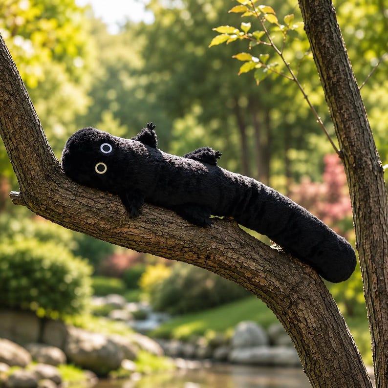 May include: A long, black plush toy shaped like a salamander or lizard with a soft, fuzzy texture. It features two prominent, circular white eyes with black pupils and four small, stubby limbs extending from its elongated, cylindrical body.