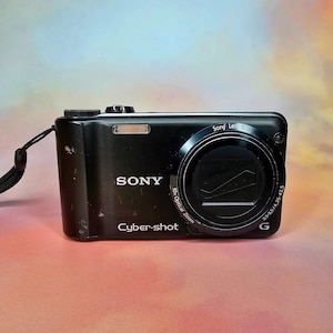 Puede incluir: Una cámara digital Sony Cyber-shot negra con un objetivo zoom óptico de 10x. La cámara tiene un botón plateado en la parte superior y el texto "Sony Lens" y "3.5-5.5/4.25-42.5" impreso en el lateral.