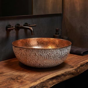 May include: A round vessel sink featuring a hammered copper interior and a textured white exterior finish. The sink is paired with a wall-mounted dark bronze faucet with two handles, positioned above a natural wood countertop with a live edge.