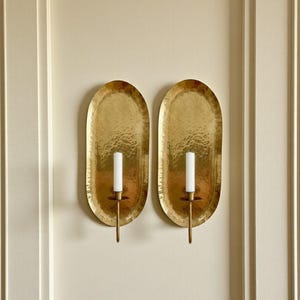 May include: Two gold-colored, oval wall sconces with a hammered texture. Each sconce holds a white candle. The sconces are mounted on a cream-colored wall with vertical trim.
