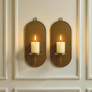 May include: Two wall-mounted candle holders with lit candles. The holders are oval-shaped with a brass finish and a small loop at the top for hanging. Each holder has a small, round platform to hold a white pillar candle. The candles are lit with a warm glow.