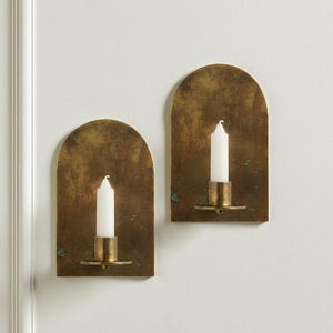 May include: Two wall-mounted candle holders with a brass finish. Each holder has an arched top and a small, round base to hold a white candle. The holders are mounted on a white wall, creating a warm and inviting ambiance.