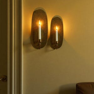 May include: Two oval-shaped wall sconces with lit candles. The sconces have a brass finish and are mounted on a beige wall. The candles are white and have a warm glow. The sconces are a decorative lighting fixture.
