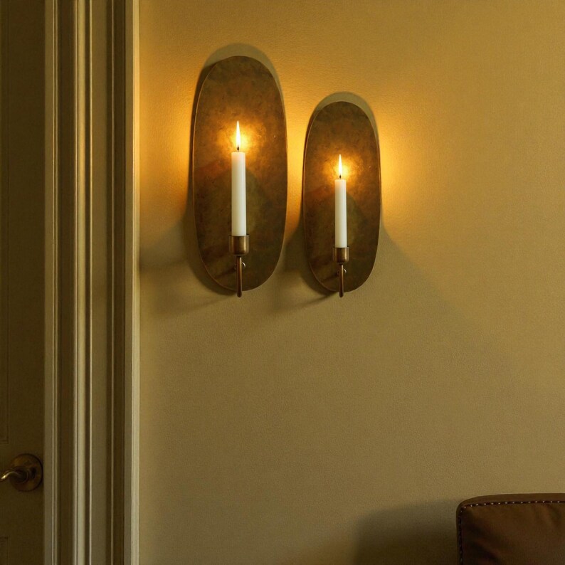 May include: Two oval-shaped wall sconces with lit candles. The sconces have a brass finish and are mounted on a beige wall. The candles are white and have a warm glow. The sconces are a decorative lighting fixture.