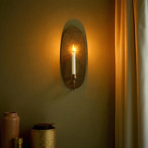 May include: A wall-mounted candle holder with a lit white candle. The holder has an oval, brass-colored backplate and a copper-toned arm. The candle flame casts a warm glow against the olive-green wall.