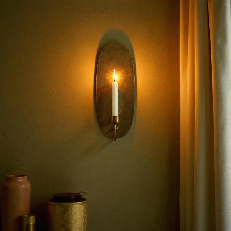 May include: A wall-mounted candle holder with a lit white candle. The holder has an oval, brass-colored backplate and a copper-toned arm. The candle flame casts a warm glow against the olive-green wall.