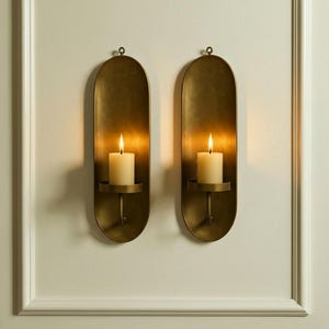 May include: Two antique brass wall sconces, each with a lit ivory candle. The sconces have an oval shape with a small loop at the top for hanging. The candles sit in a small, round holder. The sconces are mounted on a white wall.