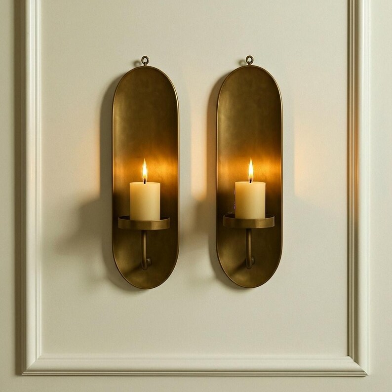 May include: Two antique brass wall sconces, each with a lit ivory candle. The sconces have an oval shape with a small loop at the top for hanging. The candles sit in a small, round holder. The sconces are mounted on a white wall.