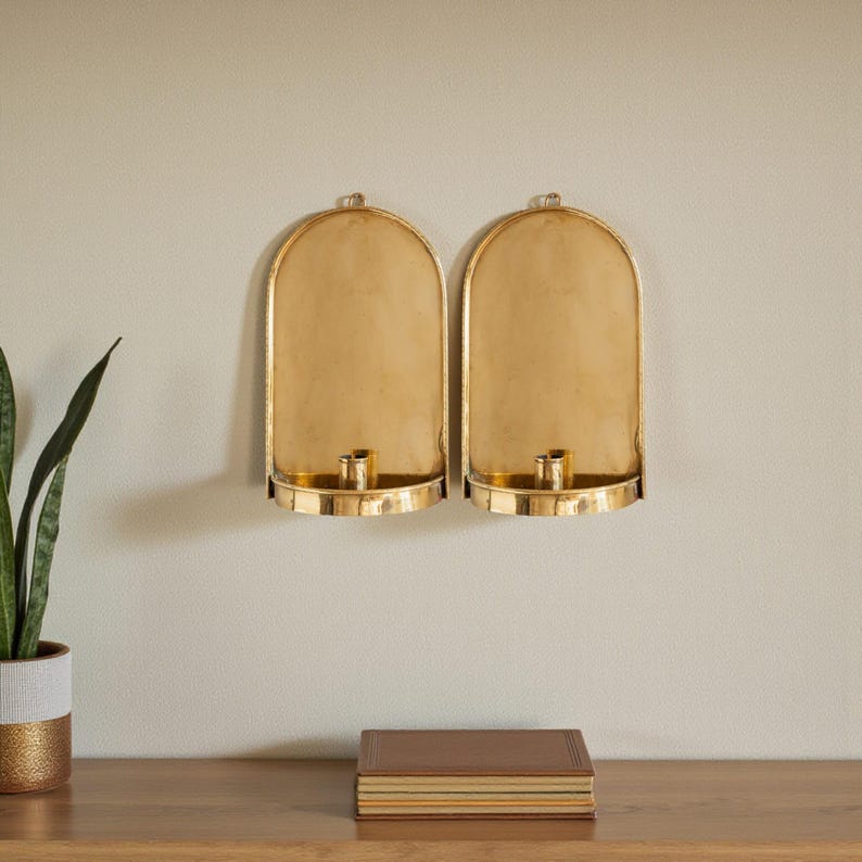 Peut inclure: Deux appliques murales dor&eacute;es avec un porte-bougie &agrave; la base. Les appliques ont un design en forme d'arche et sont mont&eacute;es sur un mur de couleur claire. Une plante en pot et un livre sont sur une surface en bois en dessous.