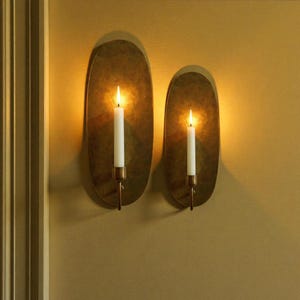 May include: Two wall sconces with lit candles. Each sconce has an oval, textured, brass-colored backplate. A white candle is held by a brass-colored holder. The sconces are mounted on a mustard-colored wall.
