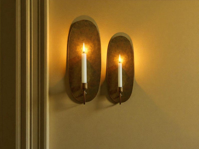 May include: Two wall sconces with lit candles. Each sconce has an oval, textured, brass-colored backplate. A white candle is held by a brass-colored holder. The sconces are mounted on a mustard-colored wall.