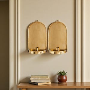 Peut inclure: Deux appliques murales dor&eacute;es avec des bougeoirs et des sommets arqu&eacute;s sont mont&eacute;es sur un mur beige. Une petite plante en pot et une pile de livres sont pos&eacute;es sur une table en bois sous les appliques.