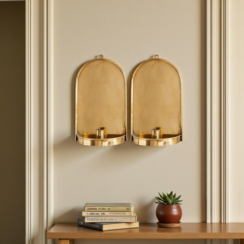 Peut inclure: Deux appliques murales dor&eacute;es avec des bougeoirs et des sommets arqu&eacute;s sont mont&eacute;es sur un mur beige. Une petite plante en pot et une pile de livres sont pos&eacute;es sur une table en bois sous les appliques.