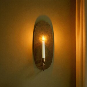 May include: A wall-mounted candle holder with a lit white candle. The holder is oval-shaped and made of a gold-colored metal. The candle flame casts a warm glow, creating a cozy ambiance. The wall is painted a warm, earthy tone.