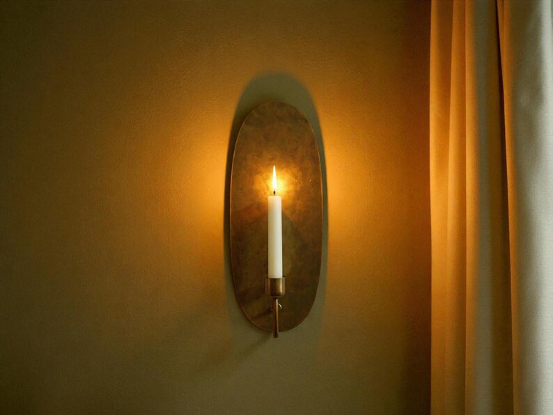 May include: A wall-mounted candle holder with a lit white candle. The holder is oval-shaped and made of a gold-colored metal. The candle flame casts a warm glow, creating a cozy ambiance. The wall is painted a warm, earthy tone.