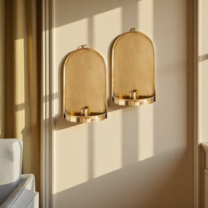 Peut inclure: Deux appliques murales dor&eacute;es avec un sommet incurv&eacute; et un petit bougeoir. Les appliques sont mont&eacute;es sur un mur cr&egrave;me, avec des ombres de la lumi&egrave;re du soleil. Les appliques sont en m&eacute;tal et ont un design simple et &eacute;l&eacute;gant.