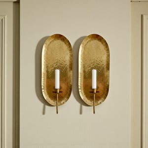 May include: Two gold-colored wall sconces with an oval, hammered metal design. Each sconce holds a white candle. The sconces are mounted on a light beige wall, adding a touch of elegance to any space.
