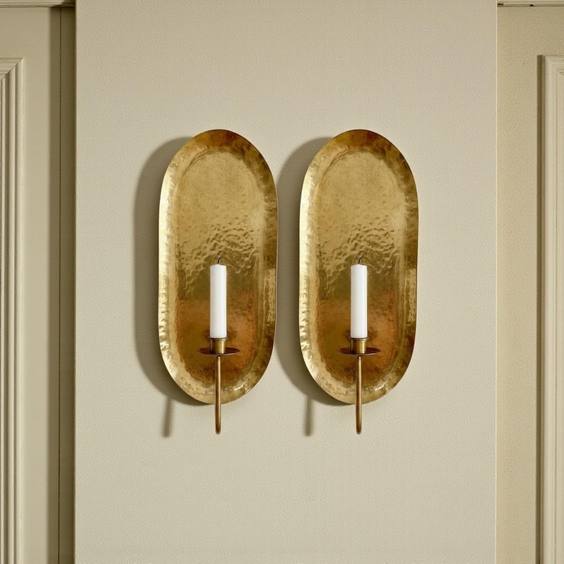 May include: Two gold-colored wall sconces with an oval, hammered metal design. Each sconce holds a white candle. The sconces are mounted on a light beige wall, adding a touch of elegance to any space.