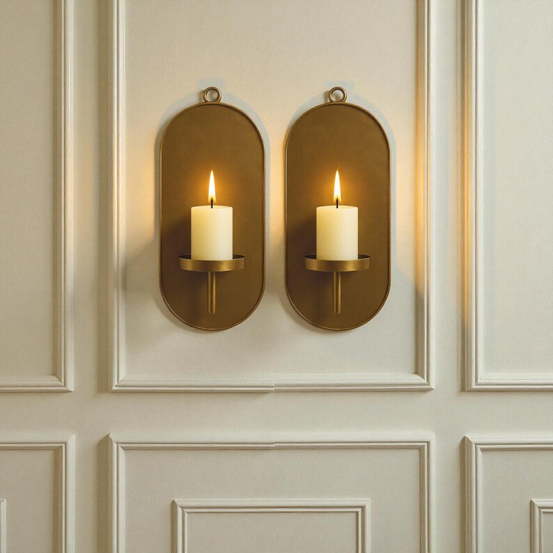 May include: Two wall-mounted candle holders with lit candles. The holders are oval-shaped with a brass finish and a small loop at the top for hanging. Each holder has a small, round platform to hold a white pillar candle. The candles are lit with a warm glow.
