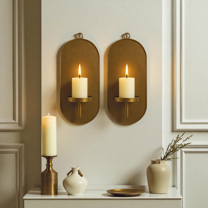 May include: Two gold-colored wall sconces with lit white candles, each mounted on an oval backplate. A third candle sits on a gold-colored candlestick. A small white vase and a gold plate are also on display.