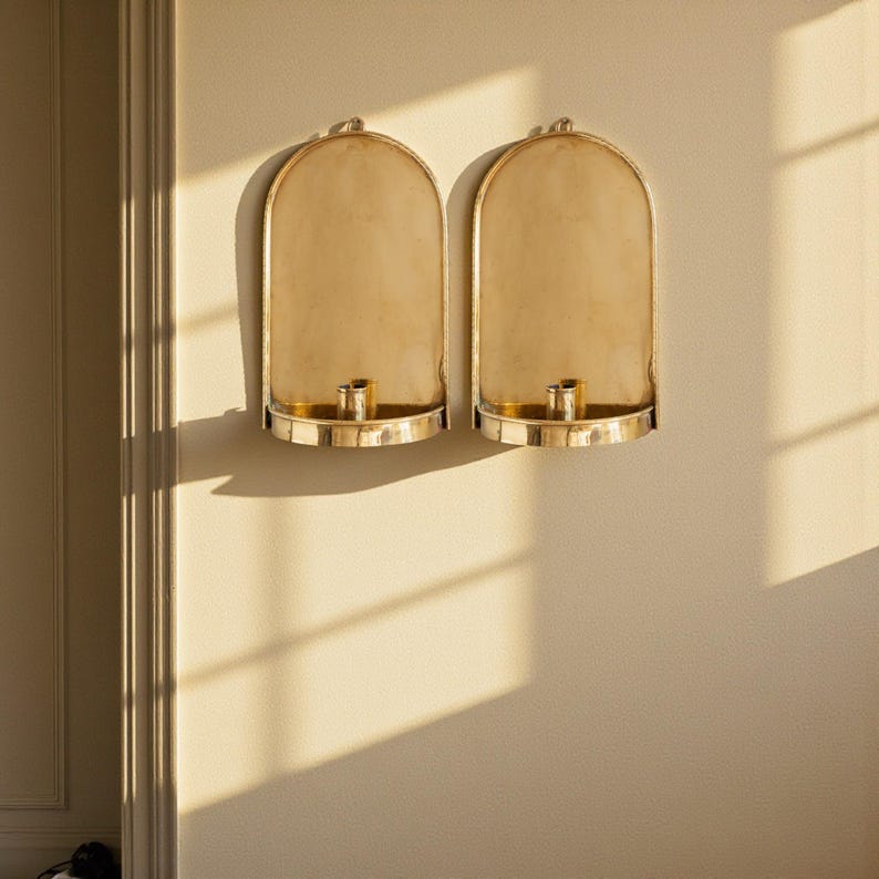 Peut inclure: Deux bougeoirs muraux dor&eacute;s. Chaque support a un dos en forme d'arche, une base circulaire et une coupelle centrale pour bougie. Les bougeoirs sont fix&eacute;s sur un mur de couleur claire, avec des ombres projet&eacute;es sur la surface.