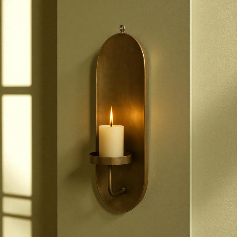 May include: A wall-mounted candle holder with a brass finish. The holder has an oval backplate and a small, round dish to hold a white pillar candle. The candle is lit, with a bright flame. The holder is hanging on a green wall.