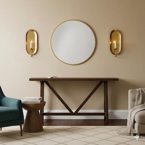 May include: A room interior featuring a large round mirror with a gold frame, a dark brown console table, and two gold sconces with lit candles. A teal armchair and a wooden side table are also visible.