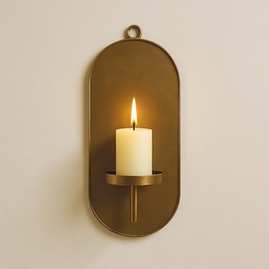 May include: A wall-mounted candle holder with a gold finish. The holder has an oval backplate with a small loop at the top for hanging. A white candle sits in a circular base, with a lit flame. The candle holder is a decorative home accent.