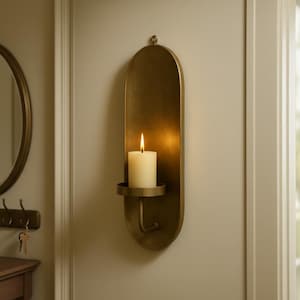 May include: A wall-mounted candle holder with a brass finish. The holder has an elongated oval shape with a small circular base to hold a white candle. A lit candle is in the center, casting a warm glow.