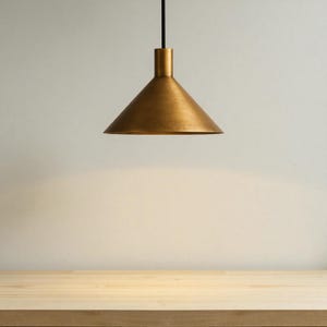K&ouml;nnte beinhalten: Eine Pendelleuchte aus Messing mit konischem Schirm, die an einem schwarzen Kabel h&auml;ngt. Das Licht befindet sich &uuml;ber einer Holzoberfl&auml;che. Das Design ist schlicht und modern, passend f&uuml;r verschiedene Einrichtungsstile.