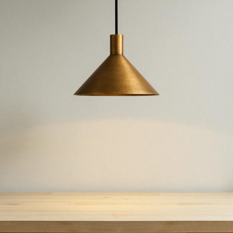 K&ouml;nnte beinhalten: Eine Pendelleuchte aus Messing mit konischem Schirm, die an einem schwarzen Kabel h&auml;ngt. Das Licht befindet sich &uuml;ber einer Holzoberfl&auml;che. Das Design ist schlicht und modern, passend f&uuml;r verschiedene Einrichtungsstile.