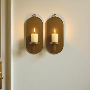 May include: Two gold-colored wall sconces with oval backplates, each holding a lit white candle. The sconces have a simple, elegant design with a loop at the top for hanging. The candles are centered on a small, round holder.