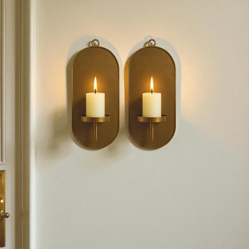 May include: Two gold-colored wall sconces with oval backplates, each holding a lit white candle. The sconces have a simple, elegant design with a loop at the top for hanging. The candles are centered on a small, round holder.