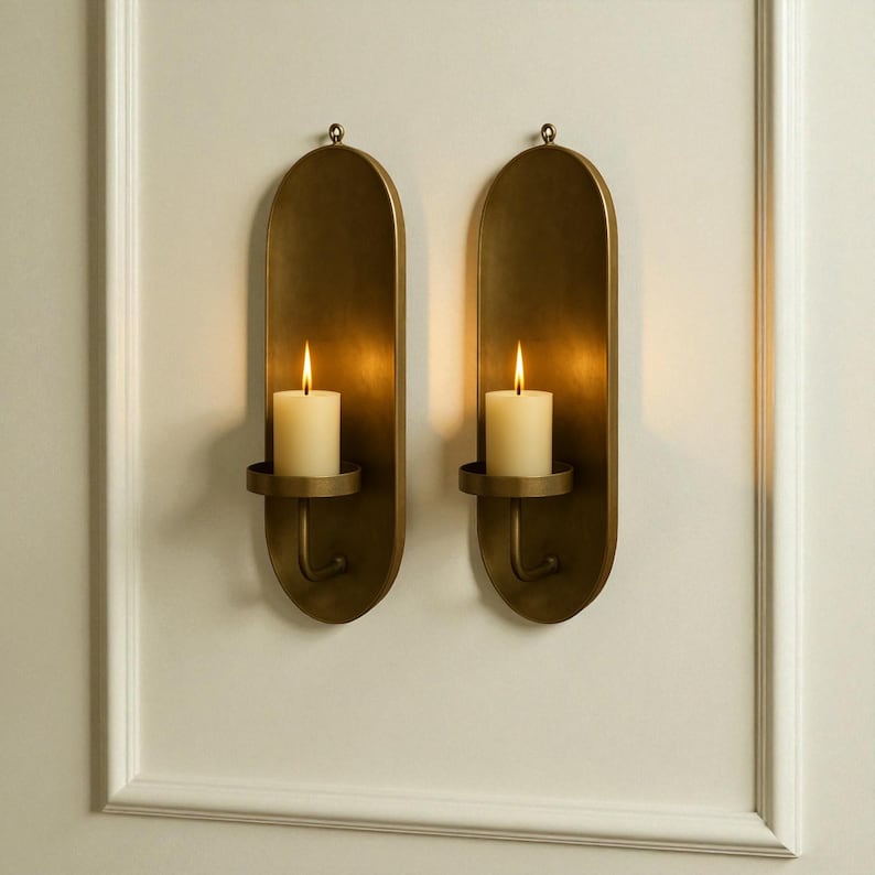 May include: Two wall-mounted candle holders with a brass finish. Each holder features an oval backplate, a circular candle cup, and a curved hook. A lit, ivory-colored candle sits in each cup, casting a warm glow. The holders are mounted on a white wall.