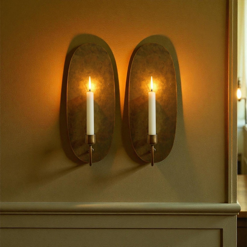 May include: Two wall sconces with oval, brass-colored backplates. Each sconce holds a lit white candle in a brass holder. The sconces are mounted on a light brown wall, creating a warm, inviting ambiance. The flames cast soft shadows.