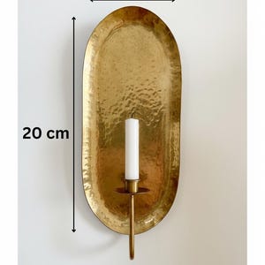 May include: A hammered brass wall sconce with a 7.9-inch height and 3.9-inch width. The sconce holds a white candle. The oval-shaped design has a textured surface and a warm, golden hue, adding a touch of elegance to any space.
