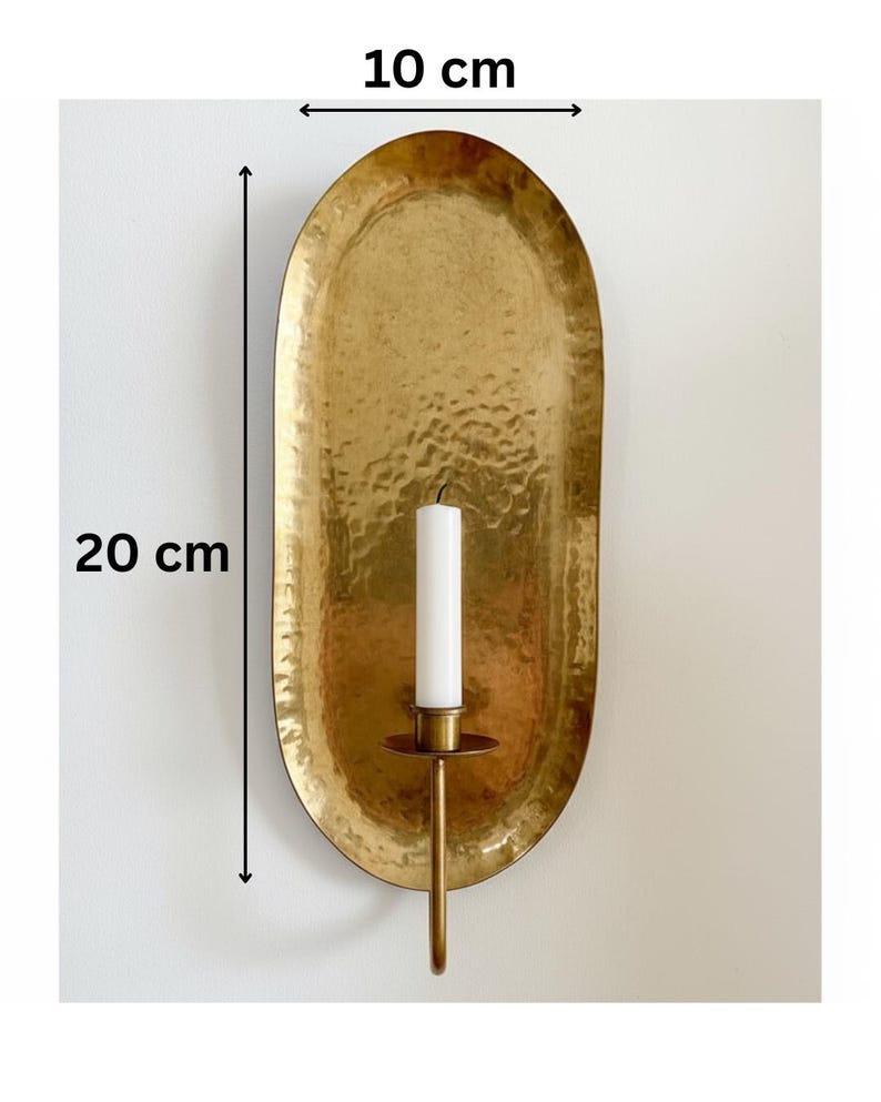 May include: A hammered brass wall sconce with a 7.9-inch height and 3.9-inch width. The sconce holds a white candle. The oval-shaped design has a textured surface and a warm, golden hue, adding a touch of elegance to any space.