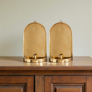 Peut inclure: Deux bougeoirs dor&eacute;s avec un sommet en forme d'arche et une base circulaire. Chaque bougeoir a une seule coupelle &agrave; bougie et une petite boucle en haut. Les bougeoirs sont expos&eacute;s sur une armoire en bois fonc&eacute;.