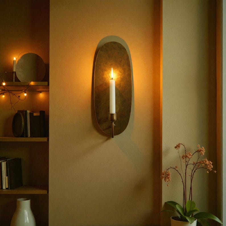 May include: A wall-mounted candle holder with a lit white candle. The holder has an oval, brass-colored backplate and a slender, brass-toned arm. The candle flame casts a warm glow, creating a cozy ambiance. The background is a muted, warm-toned wall.