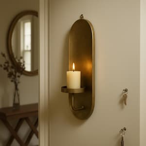 May include: A wall-mounted candle holder in a warm brass finish. The holder features an oval backplate with a small circular tray to hold a white pillar candle. A lit candle is in the holder, casting a warm glow. The holder has a hook at the top for hanging.