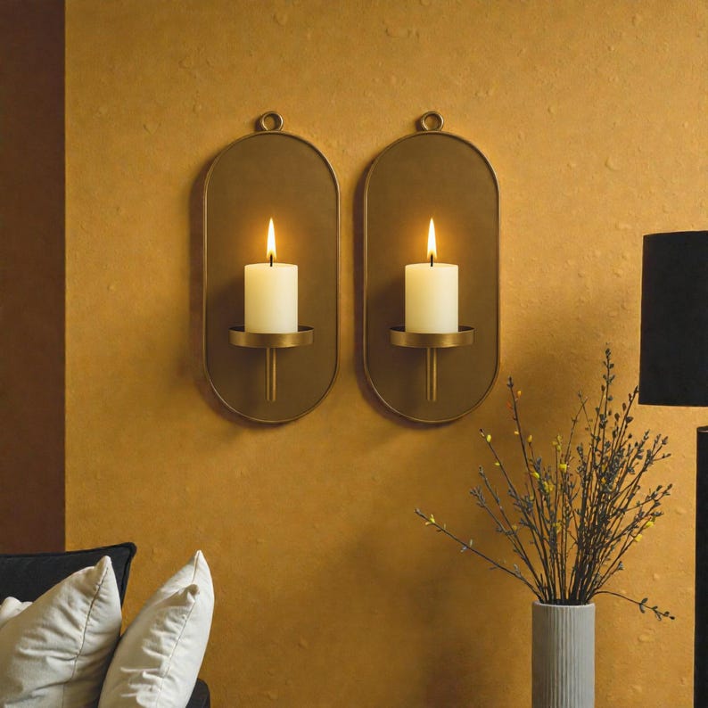 May include: Two wall-mounted candle holders with lit white candles. The holders are oval-shaped with a brass finish and a small loop at the top for hanging. The candles sit on a small, round platform. The background is a mustard-colored wall.