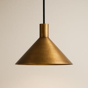 K&ouml;nnte beinhalten: Eine messingfarbene Pendelleuchte mit konischem Schirm. Das Licht h&auml;ngt an einem schwarzen Kabel und hat einen zylindrischen Aufsatz. Der Schirm hat eine glatte, metallische Oberfl&auml;che und einen breiten, offenen Boden.