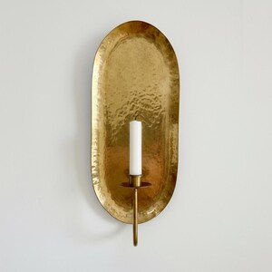 May include: A gold-colored, hammered metal wall sconce with an oval backplate. A single white candle sits in a brass holder attached to the sconce. The sconce is mounted on a white wall, creating a warm, inviting ambiance.
