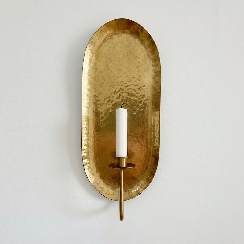 May include: A gold-colored, hammered metal wall sconce with an oval backplate. A single white candle sits in a brass holder attached to the sconce. The sconce is mounted on a white wall, creating a warm, inviting ambiance.