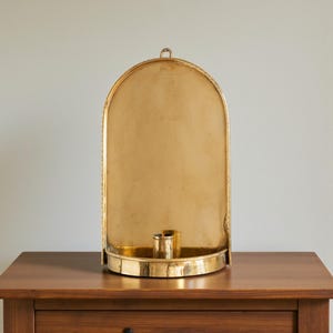 Peut inclure: Un bougeoir mural dor&eacute; en forme d'arche avec un petit porte-bougie. Le bougeoir a une bordure d&eacute;corative et une boucle en haut pour l'accrocher. Il est pos&eacute; sur une table en bois.