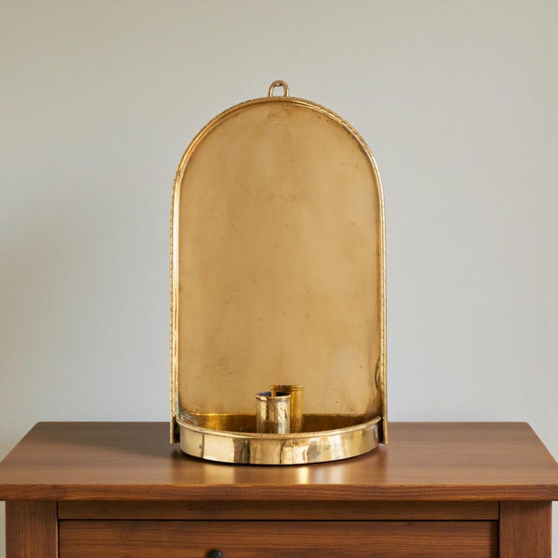 Peut inclure: Un bougeoir mural dor&eacute; en forme d'arche avec un petit porte-bougie. Le bougeoir a une bordure d&eacute;corative et une boucle en haut pour l'accrocher. Il est pos&eacute; sur une table en bois.